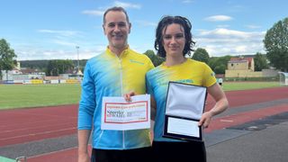 Ein Mann und eine Frau stehen auf einer Laufbahn, lächeln und halten Auszeichnungen. Der Mann hält ein Papier mit einem 'Sportlerwahl'-Logo und die Frau hält eine gerahmte Auszeichnung. Sie tragen passende gelbe und blaue Trikots mit Logos.