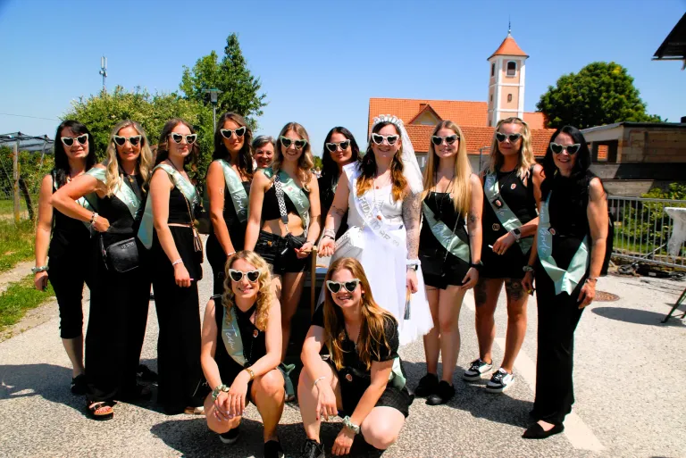 Eine Gruppe von Frauen in schwarzen und grünen Schärpen steht vor einem Gebäude mit rotem Dach. Einige knien und lächeln, während andere gerade stehen und für ein Foto posieren.