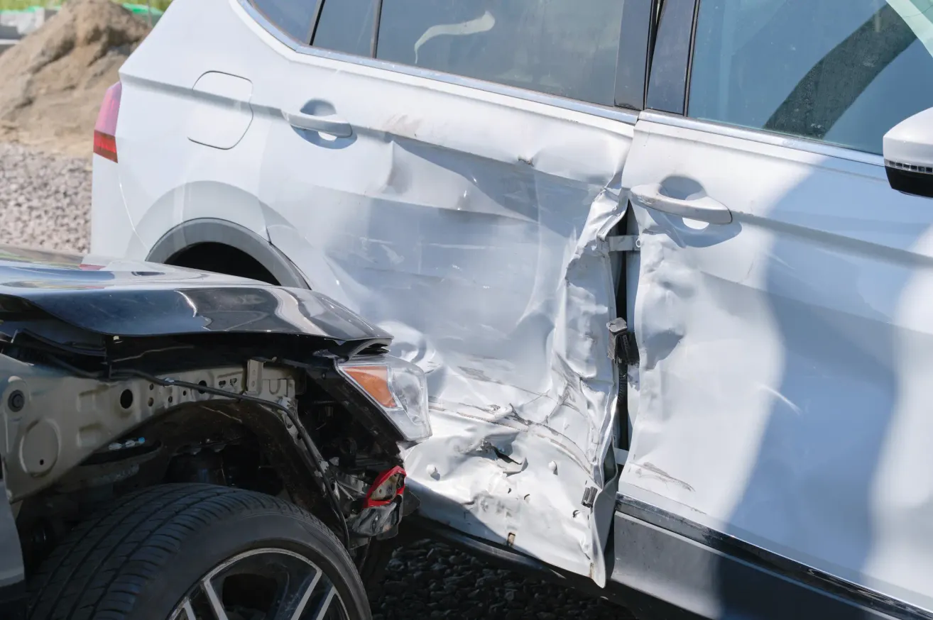 Das Bild zeigt ein weißes Auto mit erheblichen Schäden an der Seite, wahrscheinlich durch eine Kollision. Die Tür ist verbogen und der Stoßfänger ist abgerissen.