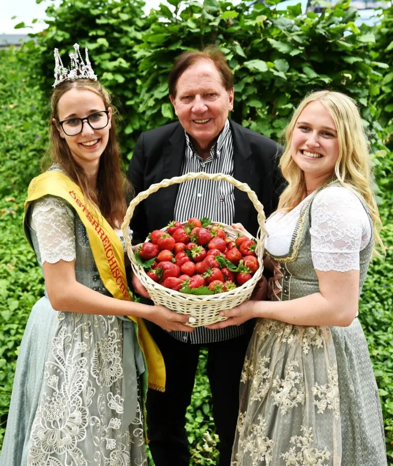 Drei Menschen, zwei Frauen und ein Mann, stehen in einem Garten. Die Frauen halten einen Korb mit Erdbeeren, und der Mann hält den Korb. Die Frauen lächeln und sind in traditioneller Kleidung gekleidet.