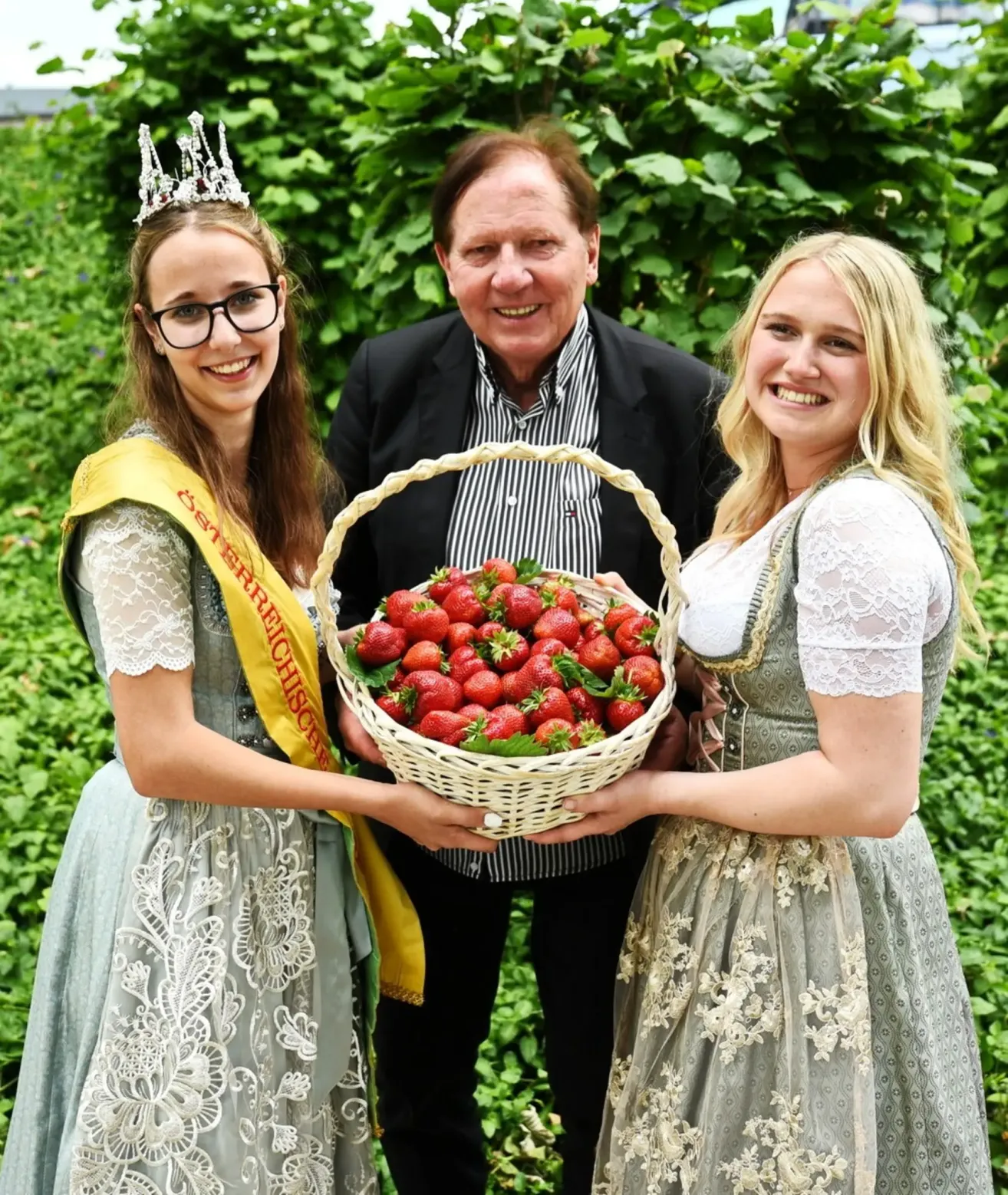Drei Menschen, zwei Frauen und ein Mann, stehen in einem Garten. Die Frauen halten einen Korb mit Erdbeeren, und der Mann hält den Korb. Die Frauen lächeln und sind in traditioneller Kleidung gekleidet.