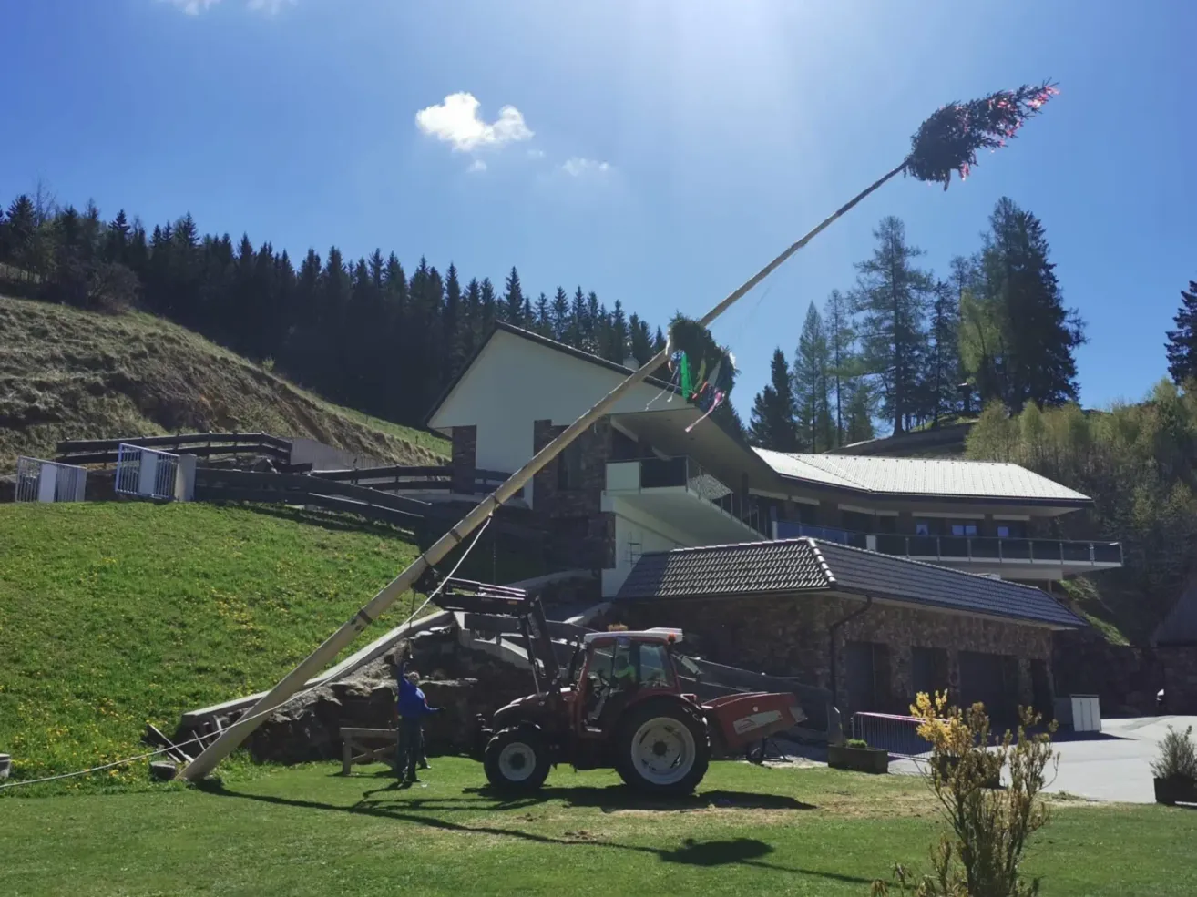 Eine Person steht neben einem Traktor und hält einen langen Pfahl mit Dekorationen an der Spitze. Ein Haus mit Treppen und Geländern ist dahinter. Bäume und ein Hügel sind im Hintergrund.