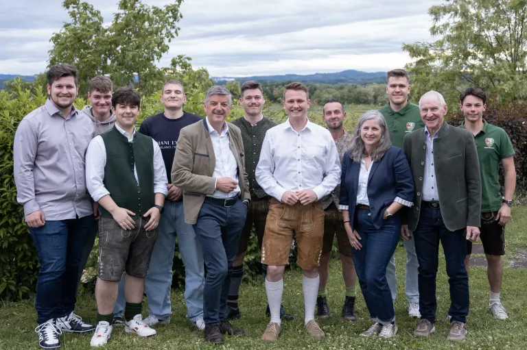 A group of people, including men and women, are standing together on a grassy field. Some are wearing traditional clothing. They are smiling and appear to be posing for a photo. Trees and mountains are in the background.