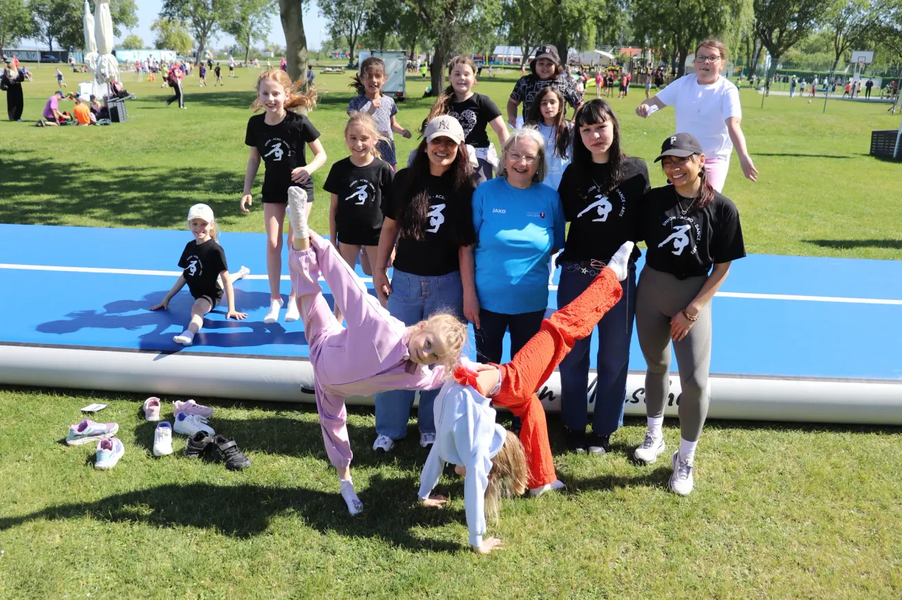 Eine Gruppe von Kindern und Erwachsenen ist im Freien auf einer blauen Matte. Sie lächeln und posieren für ein Foto, wobei einige Handstände machen.