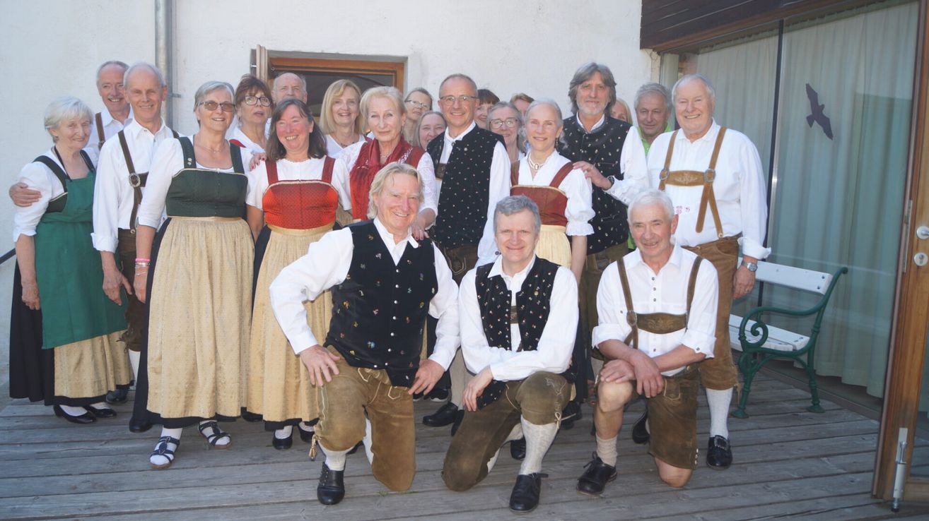 Eine Gruppe von Menschen in traditioneller bayerischer Kleidung, die lächelnd und posierend für ein Foto auf einer Holzterrasse vor einem Gebäude.