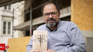 Ein Mann mit Brille und Bart hält ein Buch mit dem Titel 'Gottes Plagen' von Robert Preis. Er lächelt.