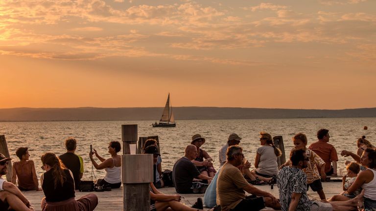 Eine Gruppe von Menschen sitzt auf einem Holzdock und genießt den Sonnenuntergang. Ein Segelboot ist in der Ferne auf dem Wasser zu sehen.