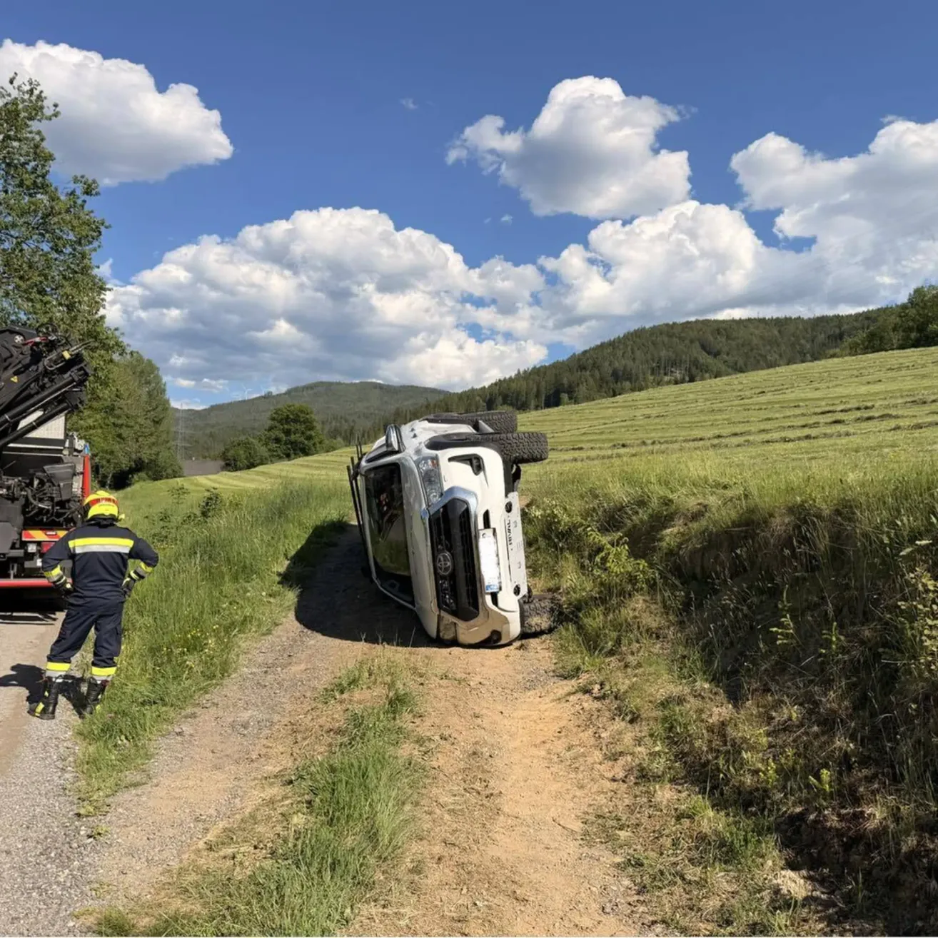 Ein weißer SUV ist auf einem Feldweg in einem Grasfeld umgekippt, ein Feuerwehrmann steht in der Nähe.