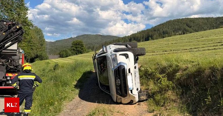 Ein umgekipptes Fahrzeug mit zwei Reifen auf dem Dach steht auf einem Feldweg in der Nähe eines Grasfeldes und einer Bergkette.