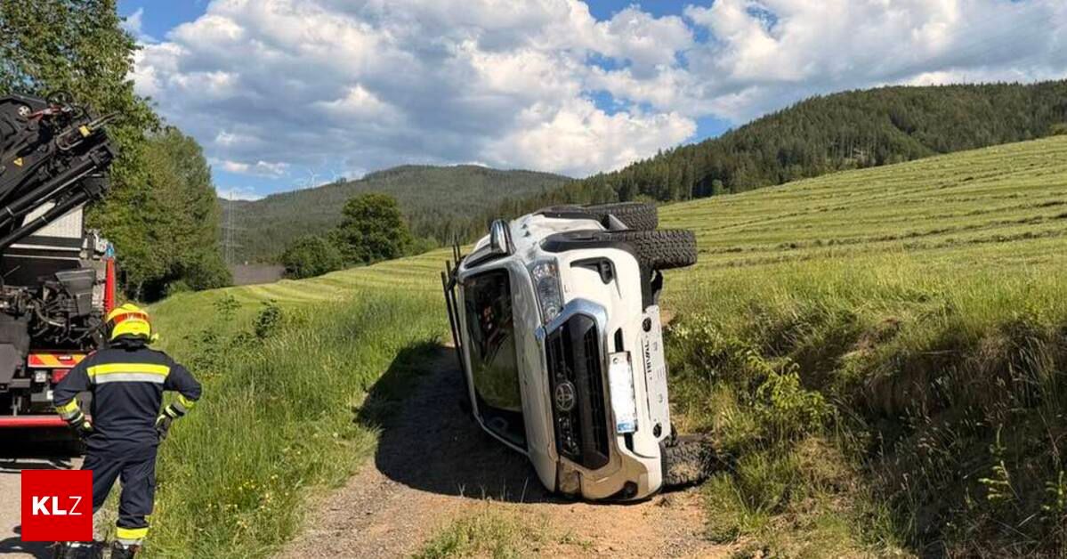 Ein umgekipptes Fahrzeug mit zwei Reifen auf dem Dach steht auf einem Feldweg in der Nähe eines Grasfeldes und einer Bergkette.