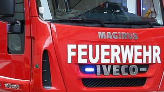 Ein roter Magirus Feuerwehrwagen mit weißen Buchstaben, die 'FEUERWEHR IVECO' und ein blaues Licht auf der Stoßstange lesen.