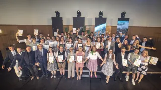 Eine Gruppe von Menschen auf einer Bühne, die Urkunden halten und für ein Foto lächeln. Sie sind in formeller Kleidung gekleidet, einige halten ihre Urkunden hoch. Scheinwerfer sind an der Wand hinter ihnen angebracht.