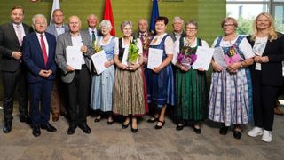 Eine Gruppe älterer Erwachsener in traditioneller Kleidung mit Urkunden und Blumen posiert für ein Foto. Hinter ihnen befinden sich Flaggen und ein grüner Hintergrund.
