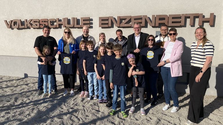 Eine Gruppe von Menschen steht vor dem Gebäude mit dem Namen 'SCHULE ENZENREI'. Sie tragen alle die gleichen T-Shirts mit einem Eulen-Design. Der Mann rechts hält ein T-Shirt und lächelt.