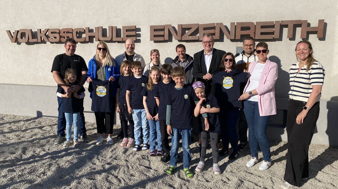 Eine Gruppe von Menschen steht vor dem Gebäude mit dem Namen 'SCHULE ENZENREI'. Sie tragen alle die gleichen T-Shirts mit einem Eulen-Design. Der Mann rechts hält ein T-Shirt und lächelt.