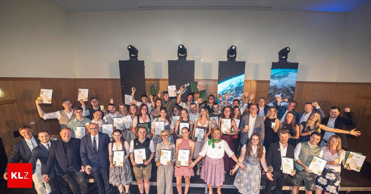 Eine Gruppe von Menschen in verschiedenen Outfits, die für ein Foto posieren und Urkunden vor einer Bühne mit großen Bildschirmen und Scheinwerfern halten.