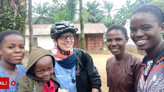 Eine Frau mit Helm und Brille posiert mit zwei Kindern in einer Dorfumgebung. Hinter ihnen steht ein Haus mit rotem Dach und Bäumen.
