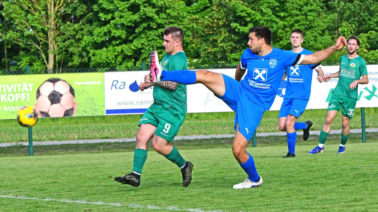 Zwei Fußballspieler spielen; einer in Grün tritt den Ball, der andere in Blau versucht zu blocken. Sie sind auf einem Feld mit einem Banner dahinter.