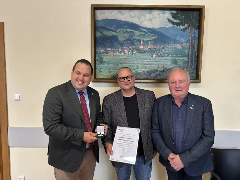 Drei Männer stehen nebeneinander und lächeln für ein Foto. Der Mann in der Mitte hält eine gerahmte Urkunde, während der Mann links eine Medaille hält. Hinter ihnen hängt ein Landschaftsgemälde an der Wand.