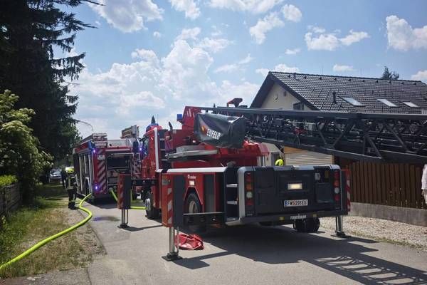 Firefighters are outside a house on a sunny day. A fire truck with its ladder extended is parked on the street. A man in a yellow vest is standing near the building.