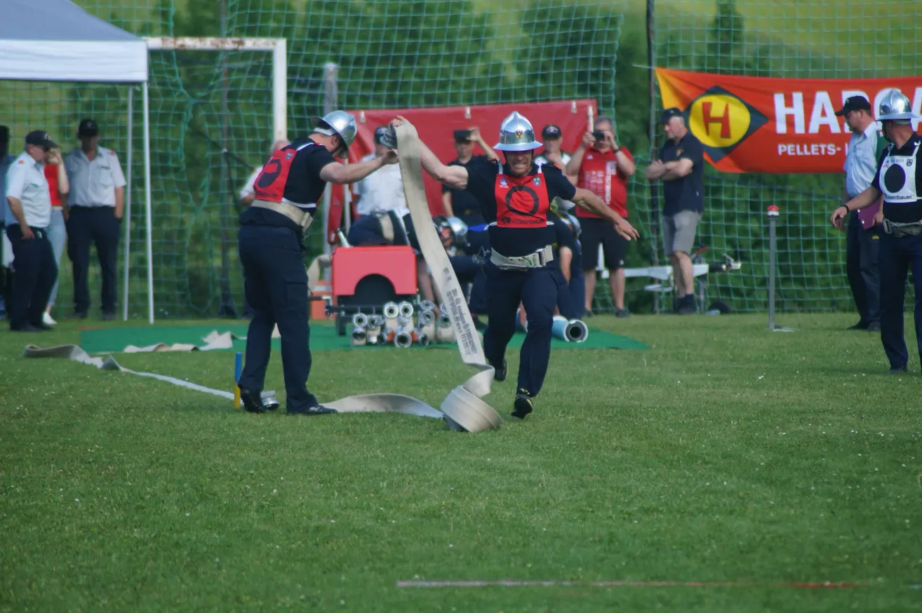 Zwei Feuerwehrleute in Uniform rennen mit einem langen Schlauch auf einem Rasenfeld. Dahinter ein rotes Tuch mit Logo und Zuschauern.