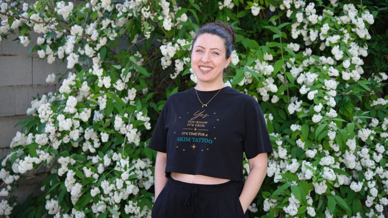 Eine lächelnde Frau in einem Crop-Top steht vor einem Busch mit weißen Blüten. Das Shirt sagt: 'Ja, du weißt, es ist wahr. Es ist Zeit für ein Skim-Tattoo.'
