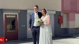 Ein lächelndes Brautpaar steht vor einem Gebäude. Der Bräutigam trägt einen Anzug und hält eine Blumenstrauß. Die Braut trägt ein Hochzeitskleid und Schleier.