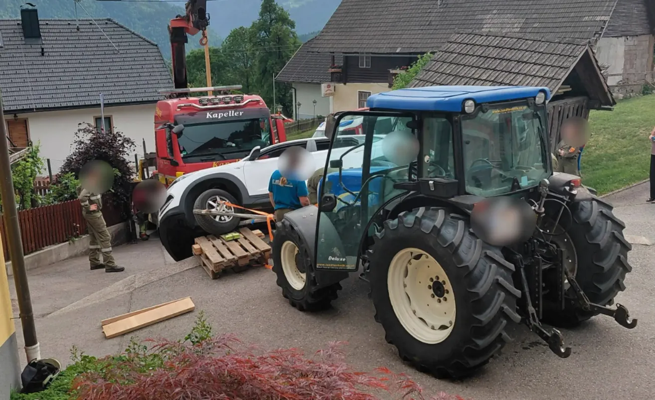 Ein Traktor steht auf der Straße, eine Person ist in der Nähe. Ein roter Feuerwehrwagen hebt einen weißen SUV an. Ein Gebäude und ein Berg sind im Hintergrund.