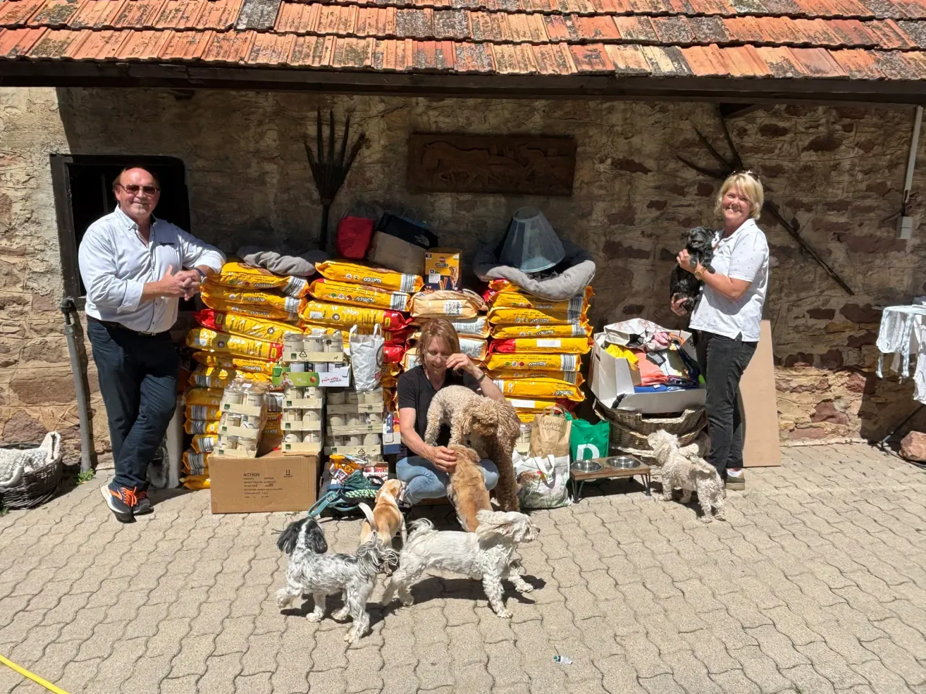 Drei Personen, zwei Frauen und ein Mann, posieren mit Hunden vor einem Steingebäude. Eine Frau hält einen Hund, während die andere einen kleinen Hund hält. Sie sind von Hundefuttertüten umgeben.