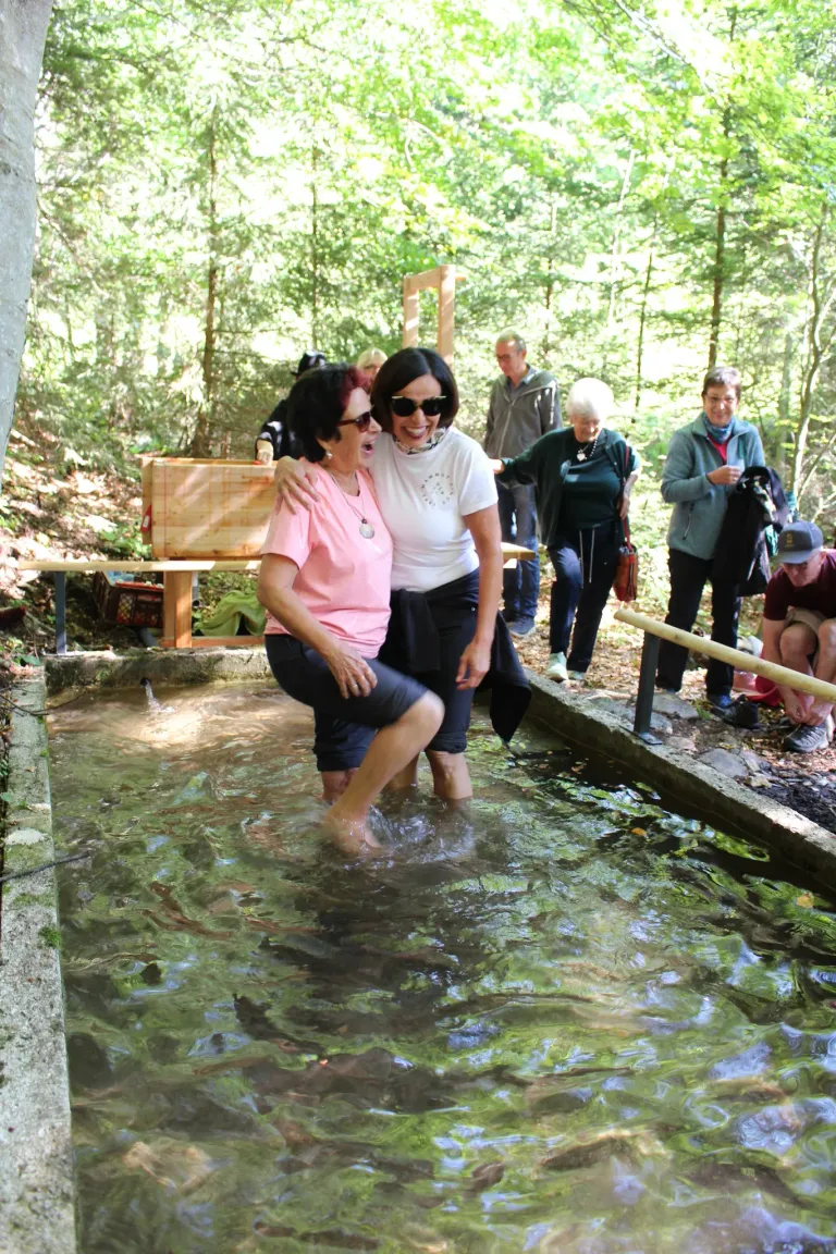 Two women are laughing and embracing in a shallow pool of water. Several people are standing around, watching them. There is a wooden bench and a wooden box nearby.