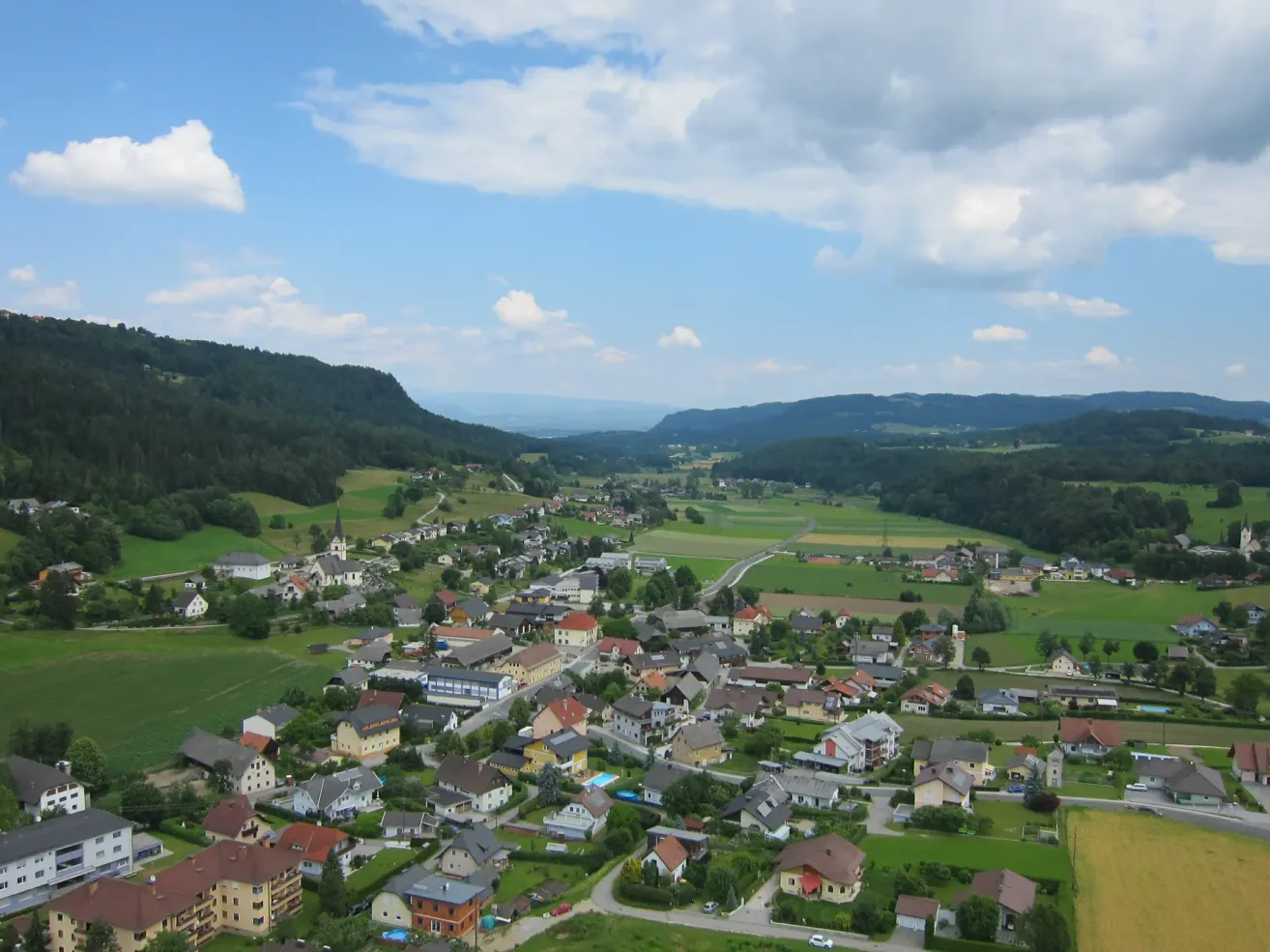 Luftaufnahme eines malerischen Dorfes in der grünen Landschaft mit Hügeln und einem blauen Himmel mit Wolken.