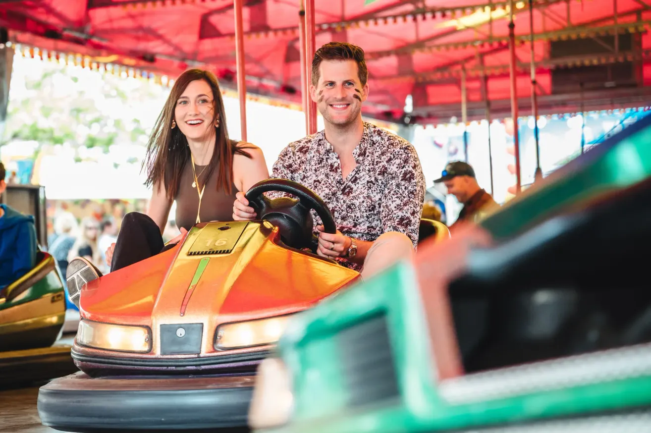 Ein Mann und eine Frau, beide mit Farbe im Gesicht, sitzen zusammen in einem Autoscooter. Sie lächeln und scheinen ihre Zeit im Vergnügungspark zu genießen.