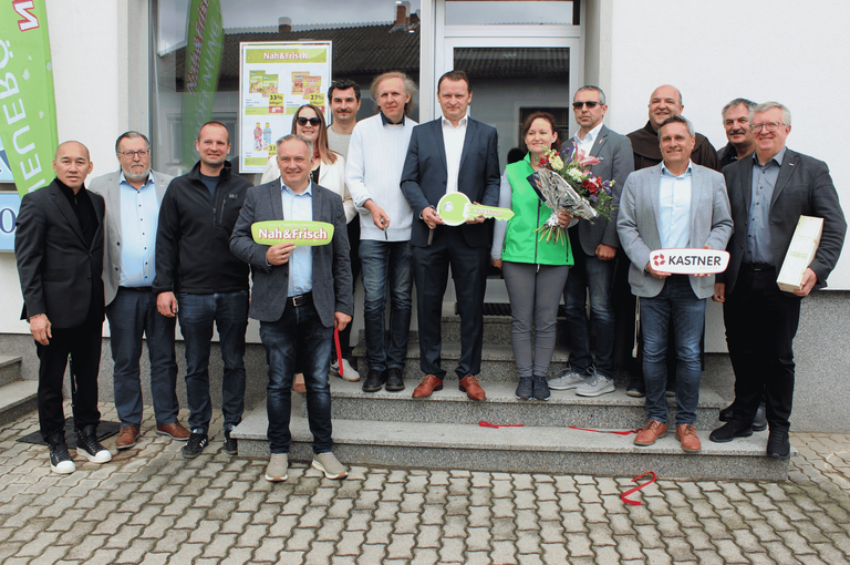 Eine Gruppe von Menschen, darunter Männer und Frauen, steht auf Stufen mit Schlüssel, Blumen und Schildern. Sie sind formell gekleidet und scheinen sich bei einer Eröffnungsveranstaltung zu befinden.