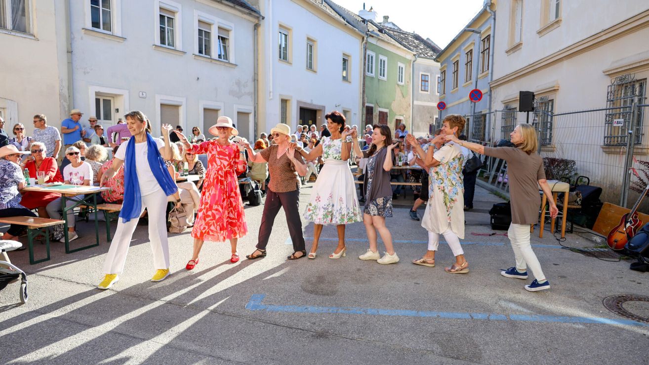 Eine Gruppe von Frauen in Sommerkleidern tanzt auf einem Stadtplatz, umgeben von Schaulustigen und Gebäuden.