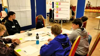 A group of young people sit around a table, possibly in a classroom, working on papers. A whiteboard with tasks and sticky notes is visible. One student holds a pen, while others appear engaged.