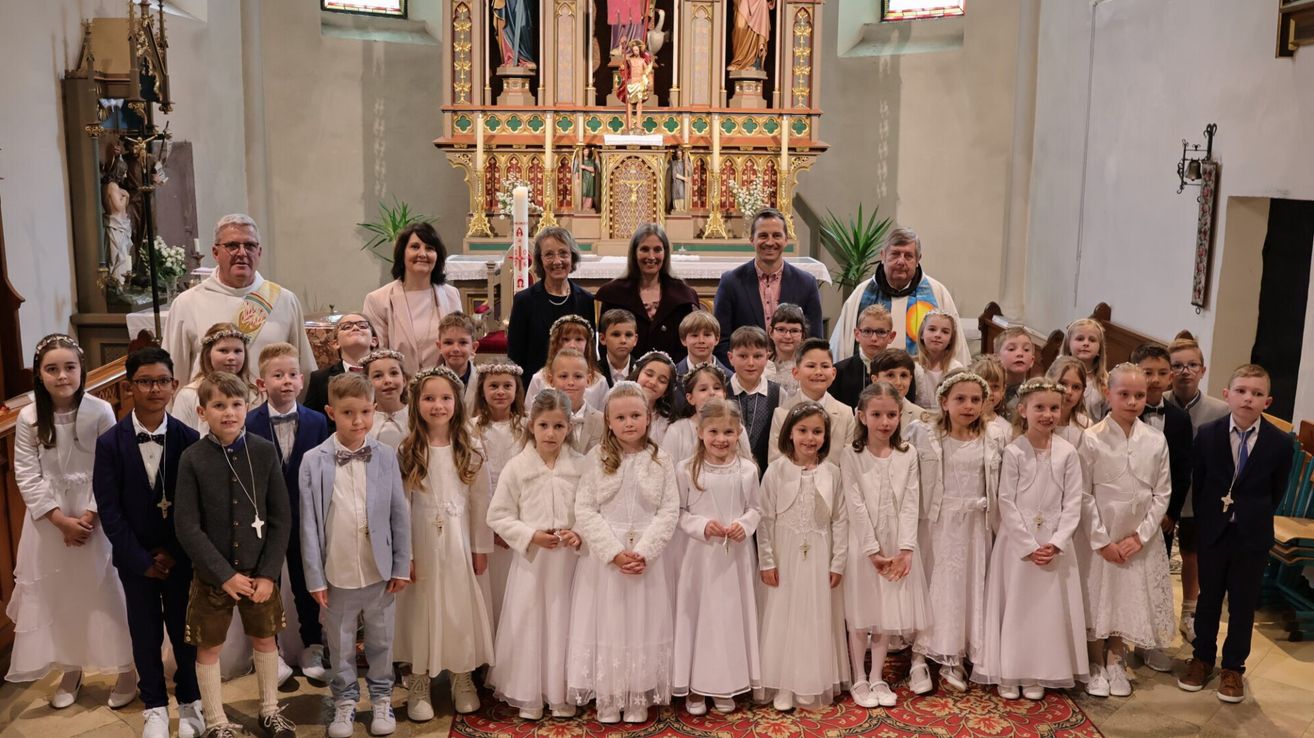 Eine Gruppe von Kindern in weißen Kleidern steht vor einem Altar, mit Erwachsenen, die hinter ihnen stehen, wahrscheinlich für eine religiöse Zeremonie.