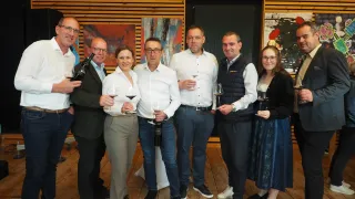 Six individuals, three men and three women, are smiling and posing for a photograph while holding wine glasses and bottles of wine in a room with wooden walls and paintings.