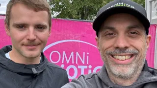 Two men smile in front of a pink backdrop with the word Omni. One man wears a gray hoodie and the other a black jacket.
