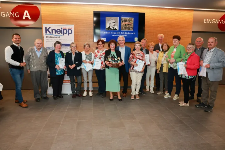 A group of individuals stand together holding certificates. Behind them, a large screen displays the words '40 Jahre Kneipp Aktiv-Club Blindenmarkt' and a picture of a man.