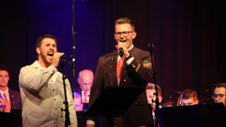 Zwei Männer in Militäruniformen singen in Mikrofone auf der Bühne. Der vordere trägt eine Brille und eine rote Krawatte, während der andere ein weißes Hemd trägt. Dahinter ist ein verschwommener Hintergrund von Menschen, die auf Stühlen sitzen.