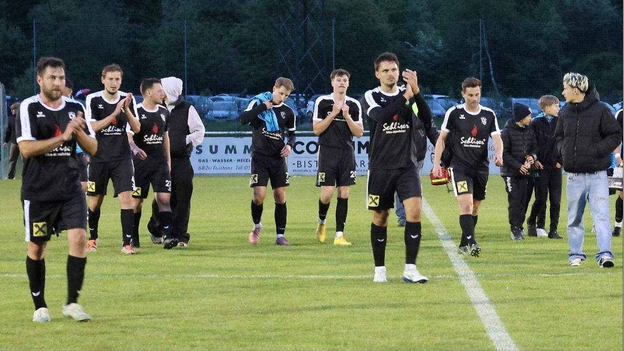 Soccer players in black uniforms are walking on a field, possibly after a match. Some wear sneakers, and one holds a red object.