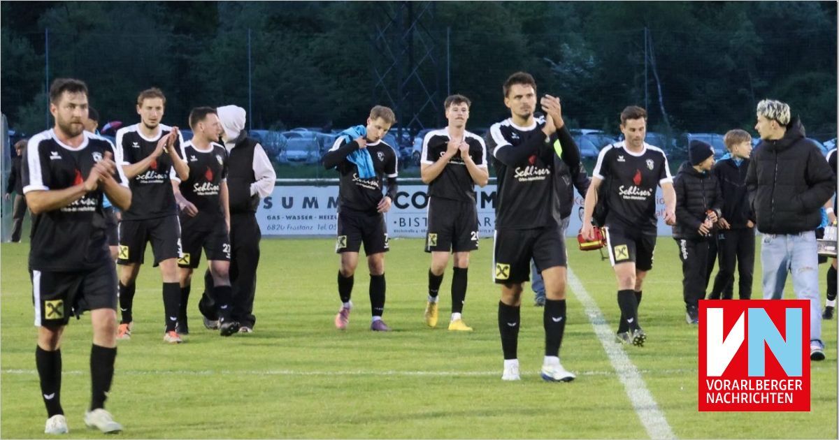 Soccer players in black uniforms walk off the field, applauding the crowd. The field has white lines and a banner with text. Trees and parked cars are in the background.