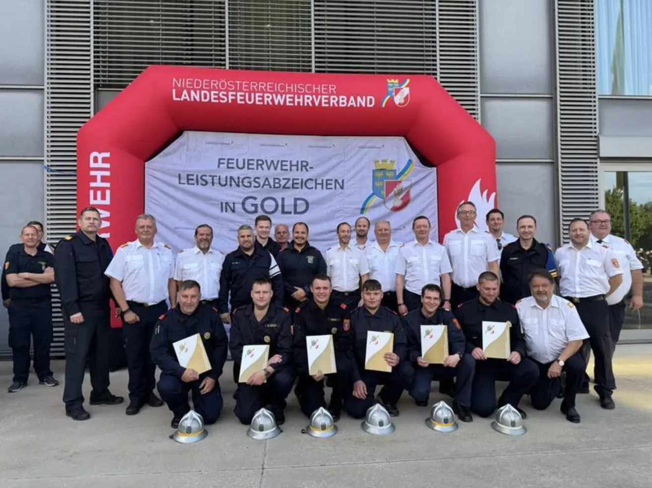 Feuerwehrleute posieren vor einem roten Bogen mit einem weißen Banner, tragen Uniformen und halten Zertifikate. Der Bogen hat Text, der 'Feuerwehr-Leistungsabzeichen in Gold' lautet.