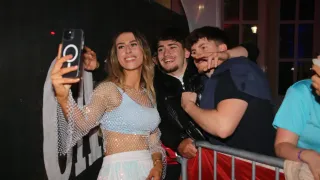 Eine Frau in einem glänzenden Kleid macht ein Selfie mit zwei Männern in einem schwarzen Mantel und einem blauen Shirt. Sie lächeln alle und stehen in der Nähe eines Zauns.