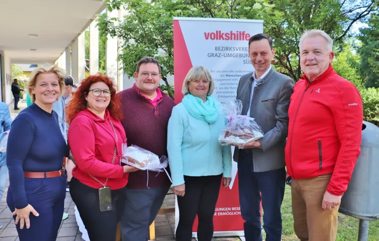 Five people are standing in front of a banner. They are smiling and holding a plastic bag with food. The banner says 'volkshilfe'. They are standing under a tree.