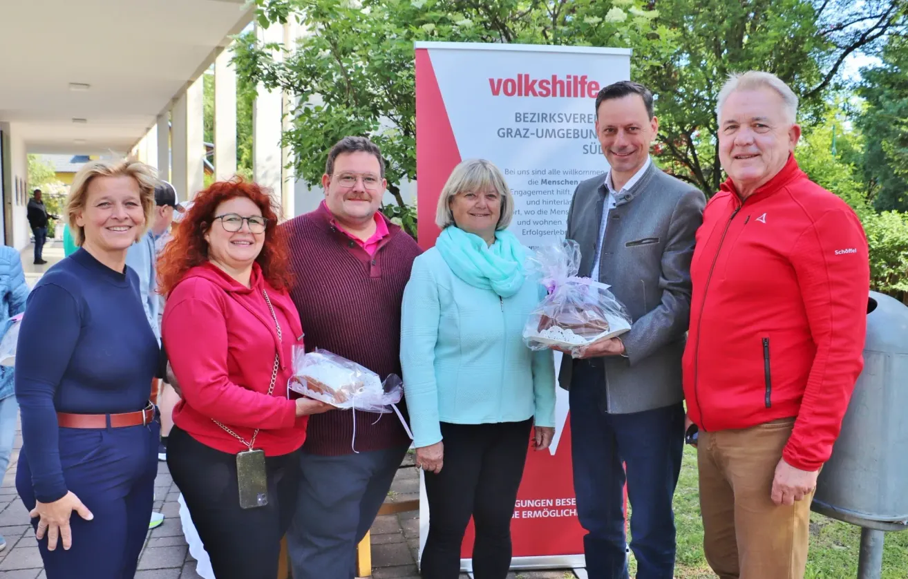 Five people are standing in front of a banner. They are smiling and holding a plastic bag with food. The banner says 'volkshilfe'. They are standing under a tree.