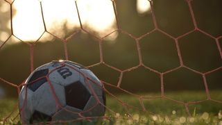 A soccer ball is caught in a red goal net on a grassy field, with the sun setting behind it.