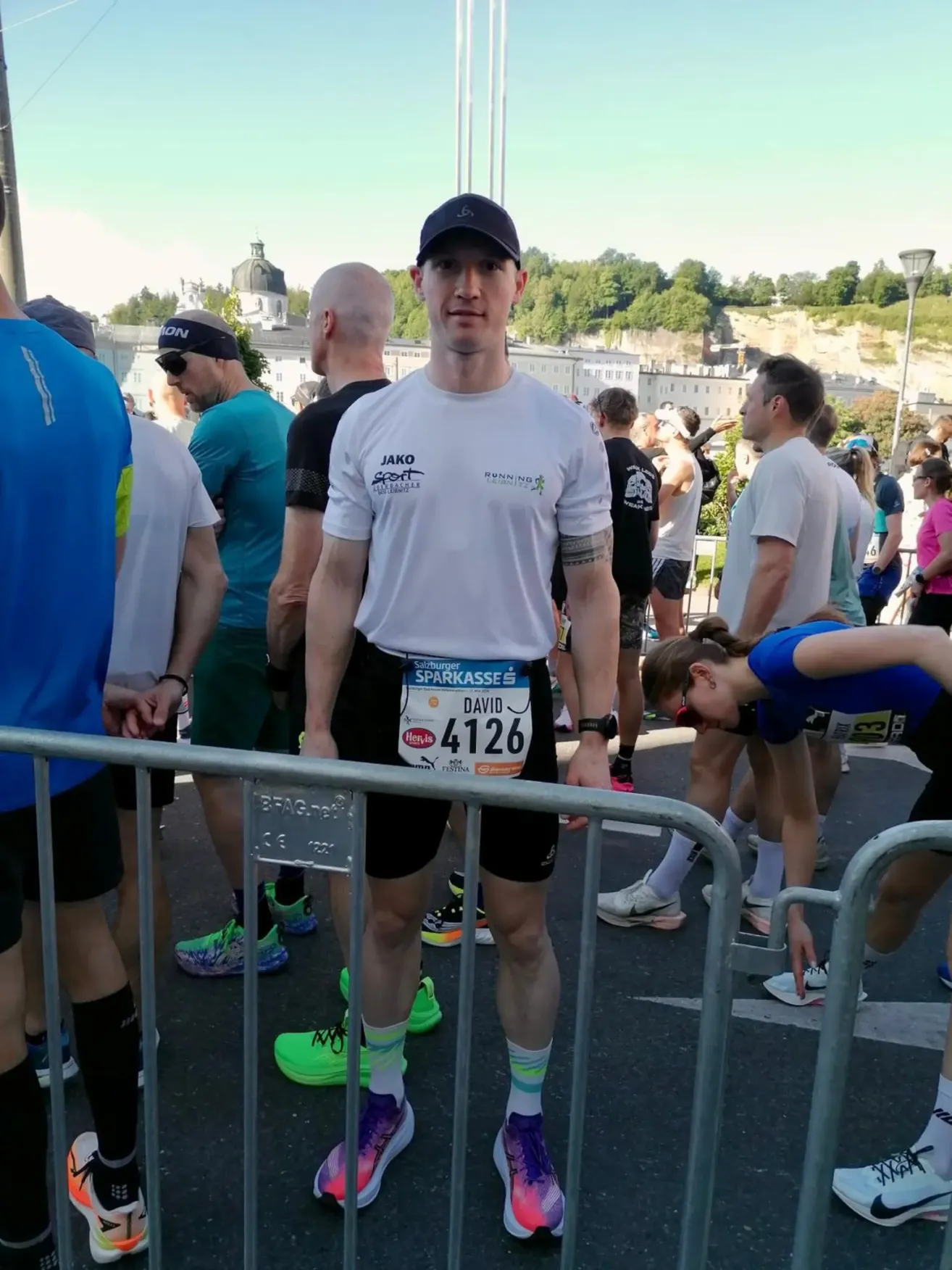 A man wearing a white shirt and black shorts stands in a crowd of runners, smiling. He has a bib with the number 4126. Behind him, a woman is bending down. Buildings and trees are in the background.
