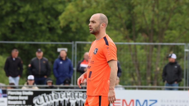 Ein Fußballspieler in einem orangefarbenen Trikot mit Logo auf der Brust schaut nach links. Hinter ihm sind verschwommene Leute, die das Spiel beobachten.