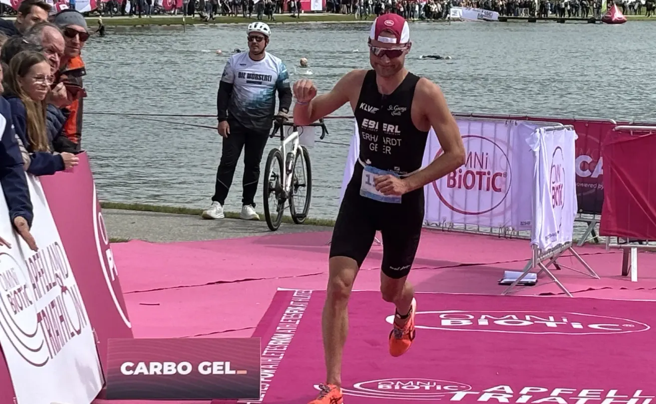 Ein männlicher Triathlet läuft auf einem rosafarbenen Weg in der Nähe eines Gewässers mit einem Fahrrad. Dahinter hält eine Person ein Fahrrad und ein weiterer Schwimmer schwimmt im Wasser.